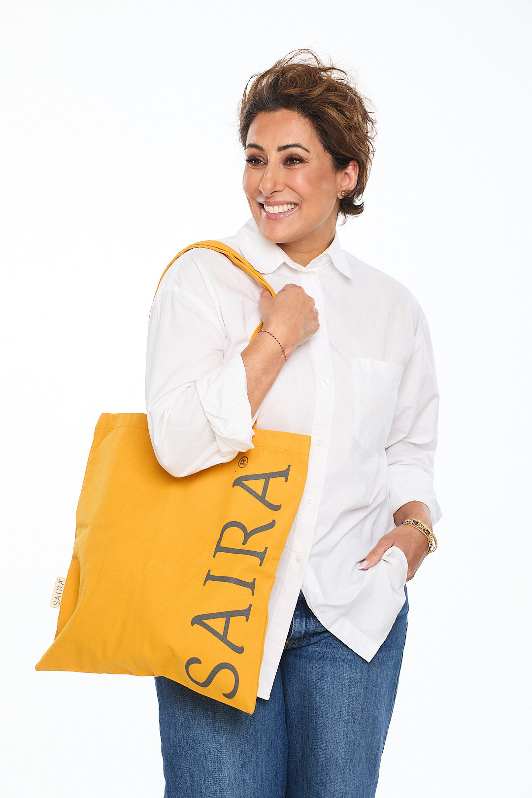 Saira Khan smiling, carrying a yellow SAIRA tote bag over her shoulder